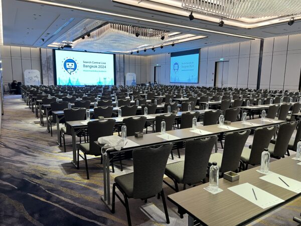 Empty conference hall with rows of tables and chairs facing large screens.