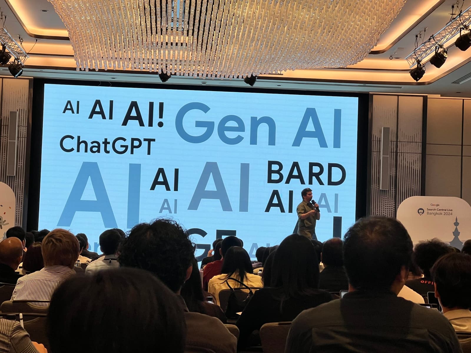 Speaker addressing a conference audience in front of a screen displaying AI-related terms.