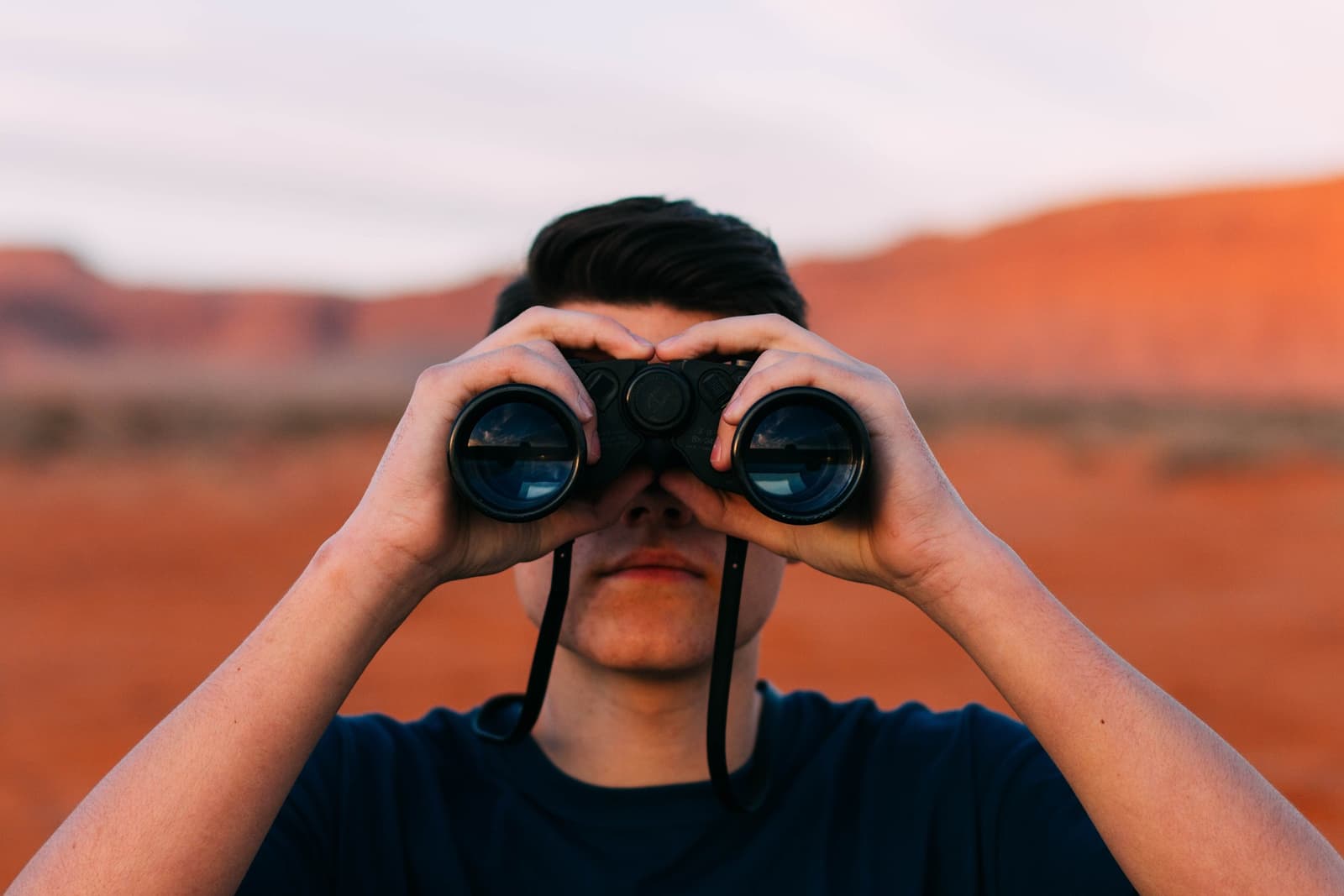 A person looking through binoculars in front of a red desert landscape.