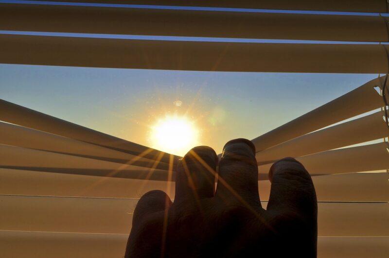 A hand parting window blinds to reveal a bright sun in the sky.