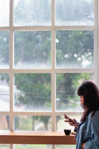 Woman looking at her smartphone beside a window on a rainy day.