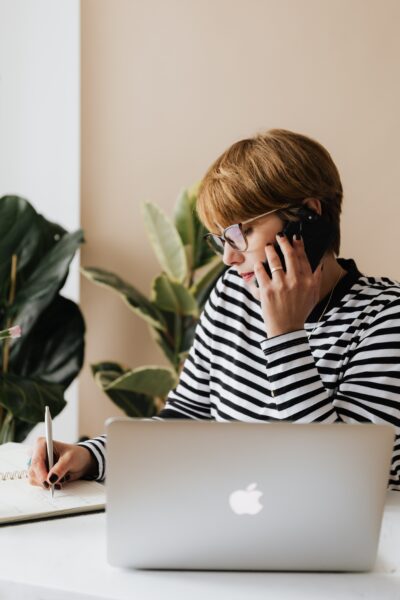 Woman talking on a phone while writing in a notebook behind a laptop.