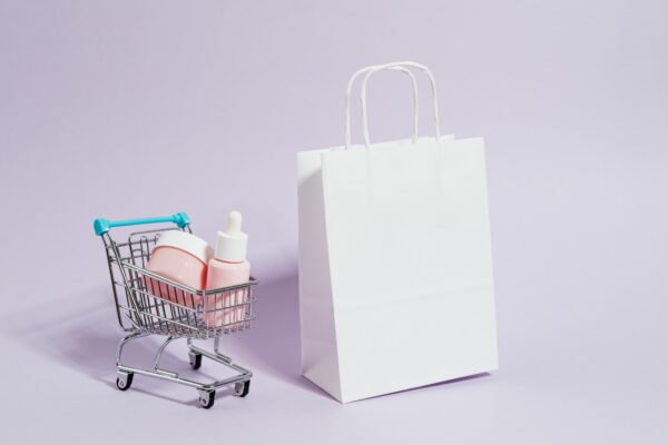 Miniature shopping trolley containing pink skincare products next to a white paper bag.